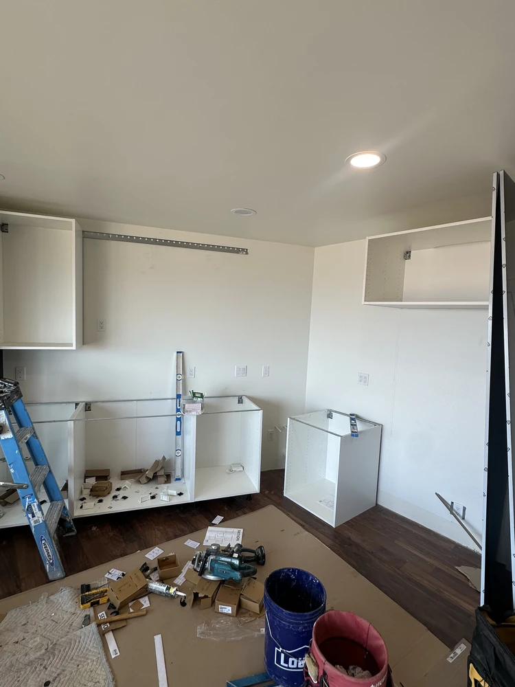Kitchen mid-remodel with cabinet boxes set, a ladder, and tools and buckets on the protected floor